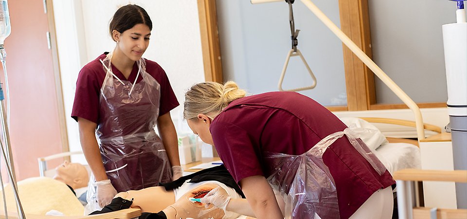 Två vårdelever står vi en sjukhussäng med en docka i. Den ena eleven rengör ett sår på benet och den andra övervakar.